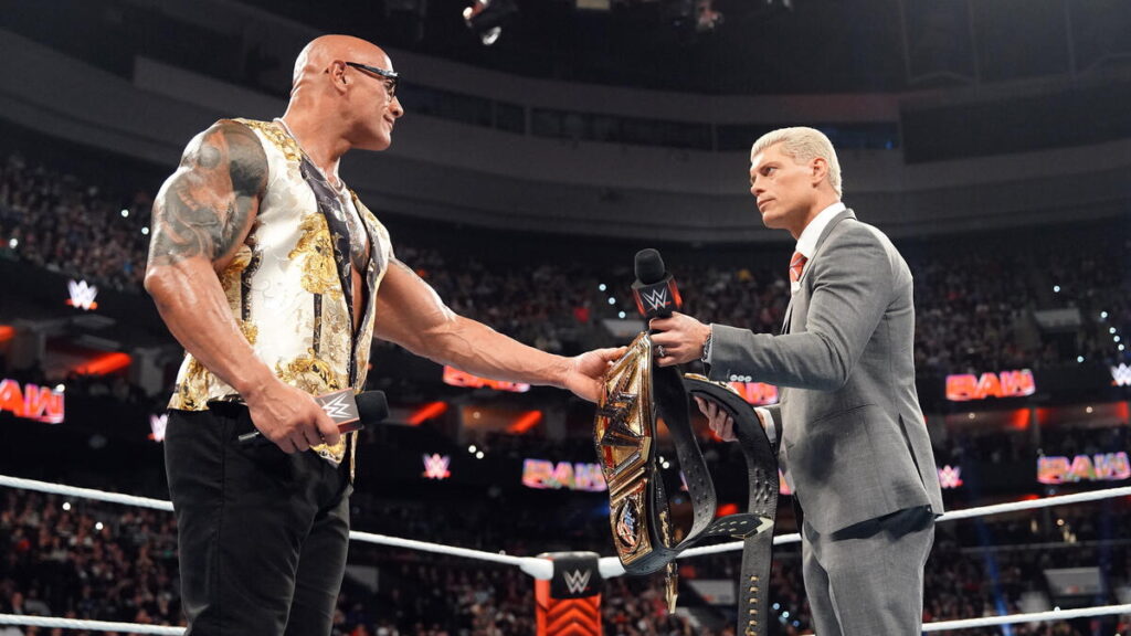 WWE star The Rock on left, exchanging championship belts to hold with Cody Rhodes on the right